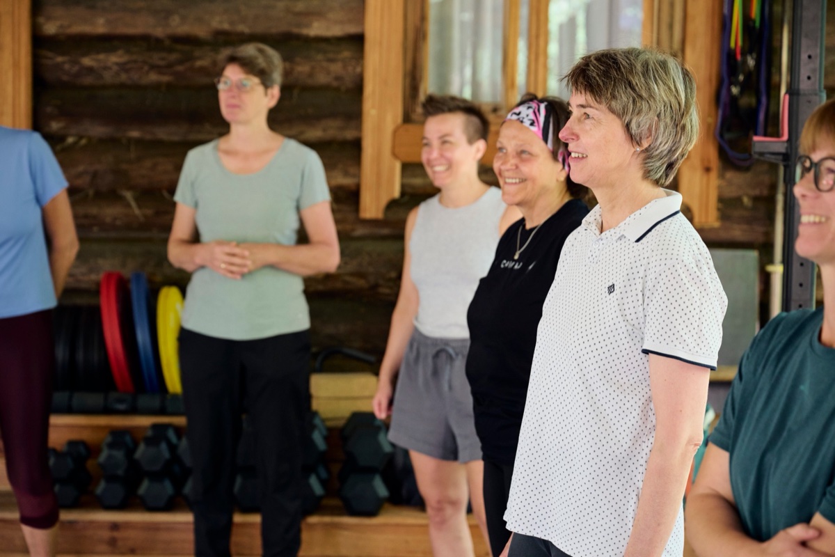 Mehrere Frauen stehen lachend im überdachten Trainingsbereich vor der Holzwand, dahinter Kurzhanteln und bunte Hantelscheiben.
