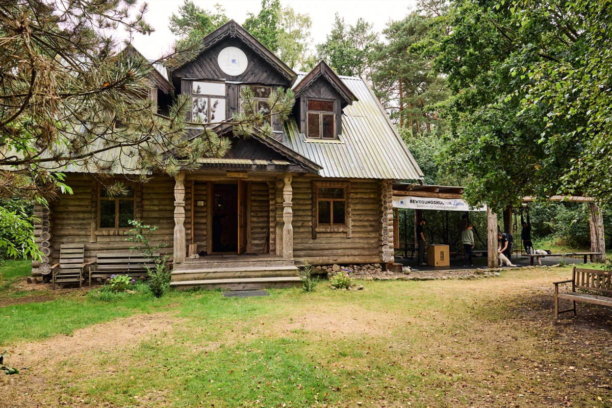 Das Trainingshaus von außen: ein Holzhaus mit Veranda auf einem Hof, umgeben von Bäumen und Wiese.